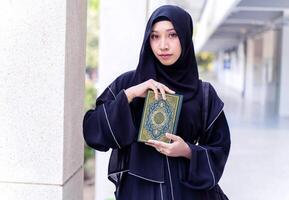 Closeup a beautiful Muslim student wearing traditional clothes portrait standing and hold Quran on walkway in her university building background. Arabic texts is meaning Glorious Quran in English. photo