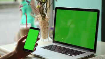 A woman using a laptop with a green screen and a smartphone in a cafe, ideal for remote work, digital nomads, or multitasking in a cozy setting. video