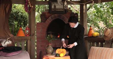 Woman in nun costume break dark chocolate into cauldron on table with Halloween decorations and pumpkin. Goth witch preparing magic treats for Halloween witchcraft in rustic gazebo with brick oven video