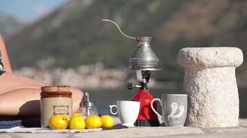 Female hands with smartphone and red camp gas burner with coffee cezve on picnic at pier in bay of Donji Stoliv, Montenegro. Coffee still life near stone bollard with blurry mountain background video