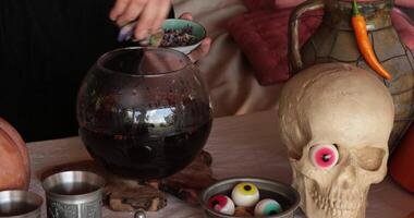 Hands make potion and put dried herbs and eyes in glass bowl on table with Halloween decorations and realistic fake skull with red candy eye. Halloween and Day of the Dead celebration props video