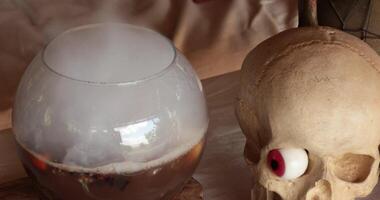 Smoke over glass bowl with Halloween decorations and realistic fake skull with red candy jelly eye. Preparing magic potion with fog made by dry ice put in cauldron. Halloween potion making concept video
