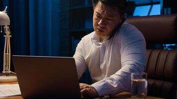 Tired angry Asian business man talking mobile phone at night evening office check documents working laptop overworked stressed Korean businessman executive manager arguing with call computer paperwork photo
