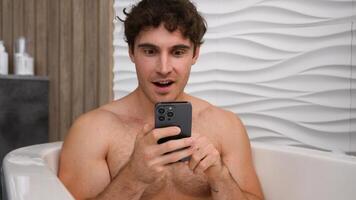 Caucasian man male guy relaxing washing in bath with soapy foam in bathtub receive offer open mouth amazed shocked surprised with smartphone reading good news in mobile phone wow astonish winning photo