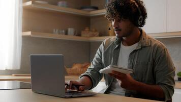 Indian man in headset look app pc browsing typing laptop write ideas questions notes review research sit at home kitchen. Arabian guy learn language improve knowledge self-education distant e-learning photo