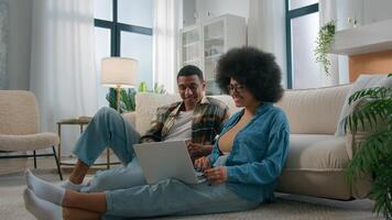 African American couple on floor at home using online laptop computer browsing shopping together pay bills choose products order delivery do high five gesture teamwork happy family in own apartment photo