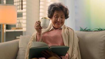 Happy smiling inspired Caucasian old woman covered with blanket on sofa drinking tea coffee with cup reading book smile atmospheric peace senior lady elderly female retired read drink at home couch photo