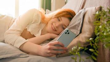 Relaxed calm smiling Caucasian girl woman lying on cozy bed at morning browsing mobile phone chatting in social media lady relaxing in bedroom use smartphone app online delivery order reading message photo