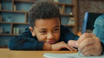 Lazy unmotivated tired exhausted fatigued little African American pupil schoolboy writing notes doing homework lying on table. Ethnic child boy kid son at desk write in copybook boring class lesson photo
