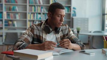 African American man writing pen notebook mobile phone smartphone call exam preparation library university guy student pupil learning study education communication gadget technology books talk male video