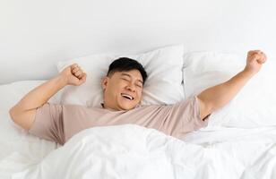 Top view of smiling asian middle aged man enjoying happy morning, lying in comfy bed with closed eyes at home, stretching his body after waking up, feeling excited and well-rested, copy space photo
