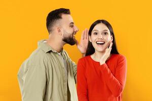 A young, Caucasian couple enjoys a playful interaction with the man whispering to the woman, who responds with a surprised, happy expression, creating a delightful, intimate atmosphere. photo