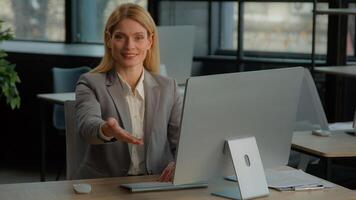 Caucasian middle-aged woman HR manager recruiter leader female businesswoman at table working on computer making inviting gesture offer to sit interview invitation consultation motivation sitting down photo