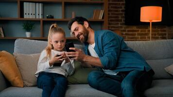 Caucasian man and little kid girl sit together at home hold modern phone browsing media content play games online adult dad teach child daughter use mobile apps showing funny on wireless device photo