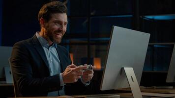 Adult businessman employee working in evening office happy male gamer Caucasian man take break playing console game on computer with gaming controller using joystick gamepad play virtual match photo