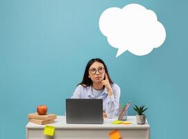 Pensive asian woman in glasses thinking with empty white speech bubble cloud with copy space, pondering questions, sitting at table isolated on blue studio background photo