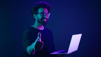 Arabian man with laptop at neon ultraviolet background Indian guy male coder programmer worker user with computer coding modern technology showing thumb up recommend online service approve good app photo