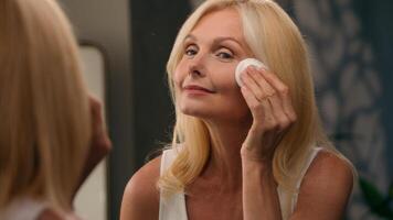 Beautiful middle-aged 50s old lady mature woman applying powder luxury cosmetics on face with cotton pad perfect smooth skin natural makeup in mirror home bathroom facial wrinkles anti-aging beauty photo