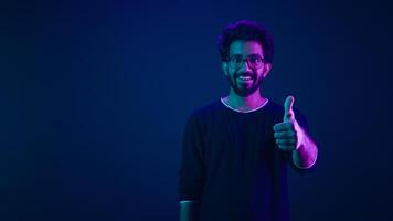 Smiling Indian man amazed Arabian IT guy Muslim male posing pointing index finger on copy space workspace isolated on neon dark blue ultraviolet background in studio showing thumb up coding promotion photo