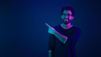 Happy Indian man showing to side on copy space Arabian guy Muslim male pointing index finger on neon ultraviolet background workspace dark blue backdrop in studio showing thumb up support recommend photo