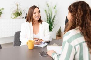 Two women are engaged in a productive conversation inside a modern office setting. One woman, dressed in a blazer, appears to be giving feedback while the other takes notes, fostering collaboration. photo