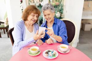 Two elderly women sit together at a round table enjoying dessert and tea. They are sharing laughter while looking at a mobile device. The cafe setting has a cozy ambiance. photo