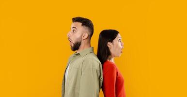 A young couple stands back-to-back with expressions of surprise, showcasing their playful connection. Bright yellow background adds to the cheerful atmosphere of togetherness and love. photo
