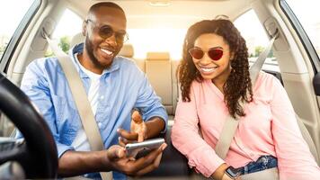 Summer Vacation Concept. Portrait of smiling African couple wearing sunglasses sitting inside luxury car, cheerful man pointing at mobile phone, showing location, using digital map application photo