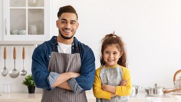 Young Arab Father And His Cute Little Daughter In Aprons Posing In Kitchen While Baking Together, Happy Dad And Small Girl Preparing Pastry At Home, Standing With Folded Arms And Smiling At Camera, photo