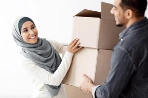 Happy lady in hijab giving her husband boxes with their stuff, moving to new apartment, closeup. Cheerful loving arab family having relocation day, packing belongings into paper boxes photo