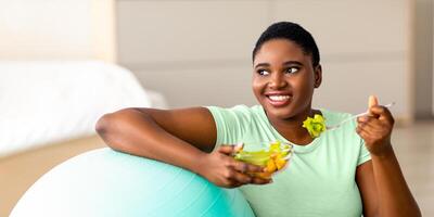 Sports and healthy nutrition for weight loss concept. Curvy black lady leaning on fitness ball, eating fresh vegetable salad at home. Plus size young woman enjoying slimming and exercising program photo