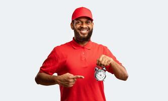 On-Time Delivery. Black Courier Pointing Finger At Clock Smiling To Camera Standing On Yellow Studio Background. photo