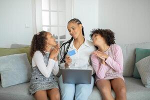 A mother enjoys a heartfelt moment with her two daughters at home. The girls express affection while the mom holds a card and sits on the couch with a laptop. photo