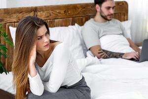 Relationship, problems, a lot of work and virtual treason. Upset woman sitting on bed and looking at husband, texting on laptop or working in modern rustic bedroom interior in evening, free space photo