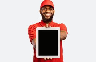 Cheerful Delivery Man Showing Digital Tablet Empty Screen Recommending Postal Courier Service Standing On Yellow Background. Mockup photo