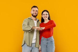 A young couple shares a joyful moment by playfully bumping their fists together. They stand in front of a bright yellow background, showcasing their affectionate connection. photo