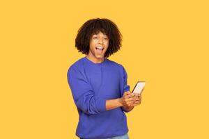 A guy with a cheerful expression holds a smartphone while standing against a bright yellow background. His casual attire reflects a modern lifestyle that embraces technology and creativity. photo