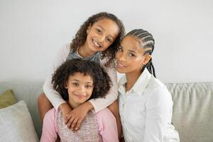 A mother shares a joyful moment with her two daughters at home. They sit together on a cozy sofa, embracing each other with smiles, highlighting their strong family bond and affection. photo