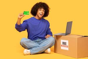 A Brazilian man sits cross-legged on the floor, holding a green card and smiling. Beside him is a delivery box with a label. A laptop is open in front of him. photo
