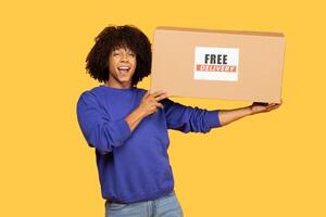 A Black man enjoying a moment of excitement holds a large cardboard box labeled free delivery. The background is bright yellow, creating a lively atmosphere as he smiles and engages with the camera. photo