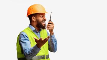 Mad african construction site supervisor yelling in two-way radio standing on white studio background. Building industry concept. Panorama, free space photo