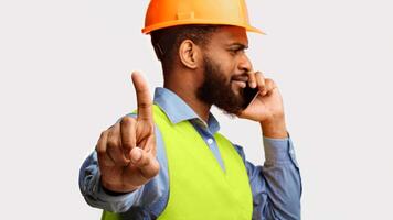 Busy Black Builder Worker Talking On Phone Gesturing Wait A Minute To Camera Standing On White Studio Background. Panorama photo