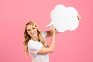 A woman with blonde hair is joyfully raising a large speech bubble while smiling. The vibrant pink background adds a playful touch to the lively atmosphere of the moment. photo