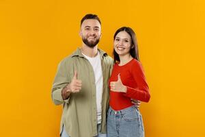 A happy young couple stands together, beaming with smiles and giving thumbs up. They pose against a vibrant yellow background, showcasing their affection and connection. photo