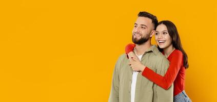 A young couple stands together, with the woman playfully hugging the man from behind. Both are smiling widely, radiating happiness in front of a bright yellow backdrop, copy space photo