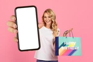 A young woman smiles brightly while holding a smartphone in one hand and a colorful birthday gift bag in the other. The background is a vibrant pink, adding to the festive atmosphere. photo