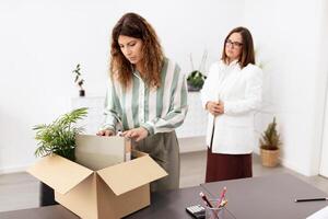 In a bright office, one businesswoman carefully sorts through a cardboard box, while another observes with a thoughtful expression. They appear to be collaborating on a workplace task. photo