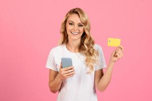 A cheerful young woman with blonde hair is standing against a bright pink background. She holds a smartphone in one hand and a yellow card in the other, exuding happiness and confidence. photo