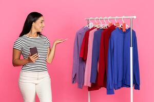 A young woman stands in front of a clothing rack, smiling as she explores colorful sweaters. The backdrop is bright pink, enhancing the lively shopping atmosphere. photo