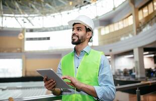 Smiling handsome young muslim male engineer with beard in hard hat and protective uniform with tablet checks project in factory or office interior. Modern gadget for professional work, copy space photo